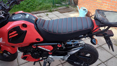 Red Honda Grom parked on a brick pavement with a garden in the background and Cheeky Seats tuck and roll cover
