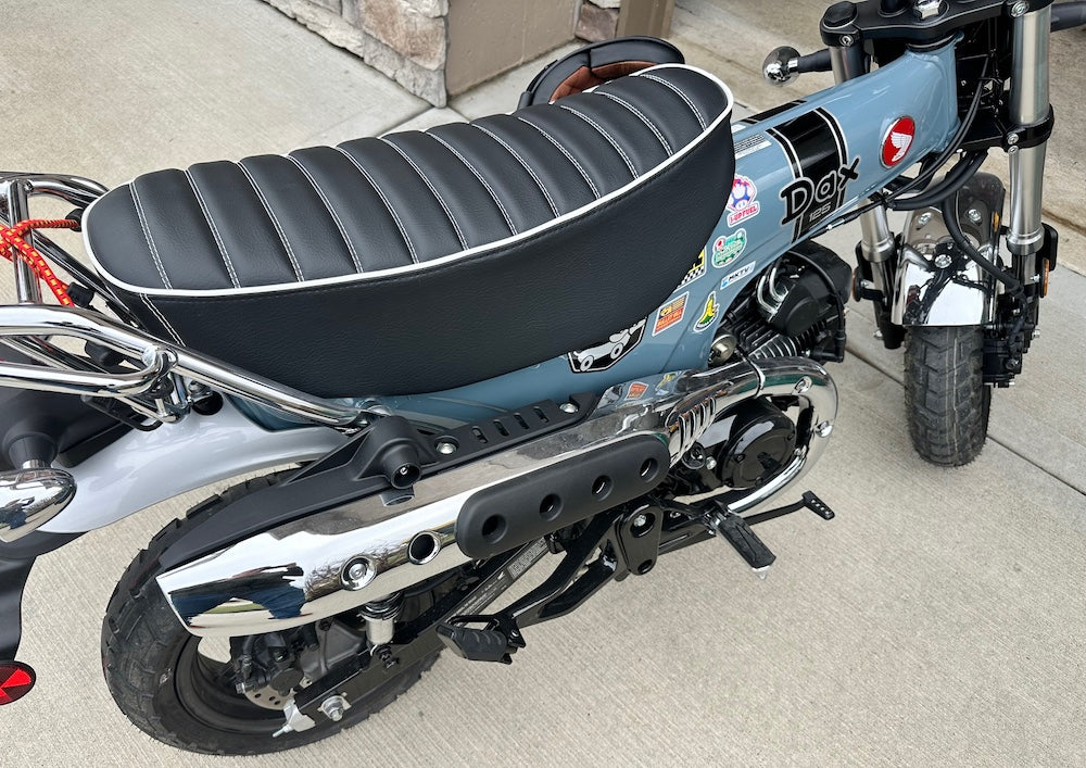 Gray-blue Honda DAX 125 with a black tuck-and-roll seat, white piping, DAX badge and red Honda wing logo, handmade by Cheeky Seats